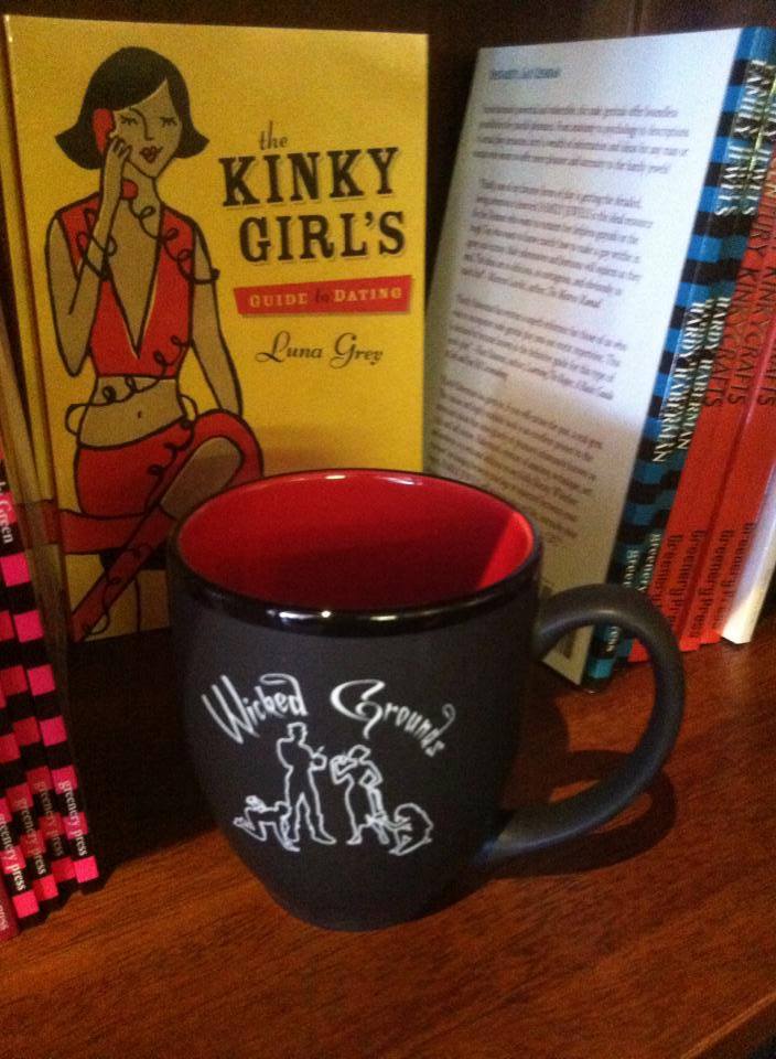 An image of a black ceramic Wicked Grounds mug directly in front of a copy of the book A Kinky Girl's Guide to Dating. 