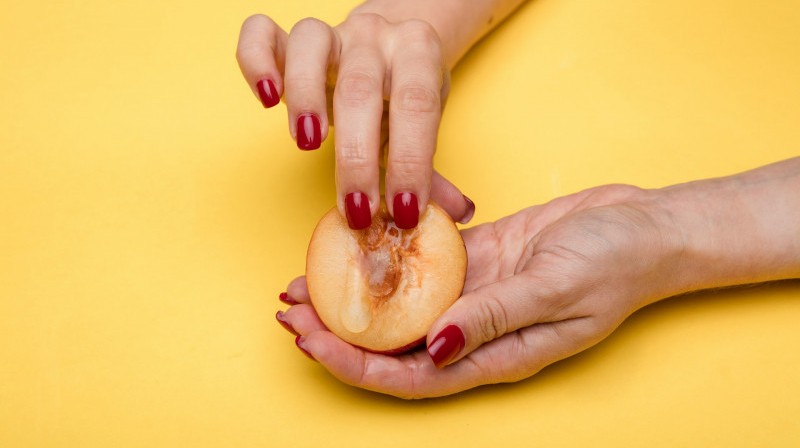 A picture of a woman putting her fingers into a ripe peach, over a yellow background.