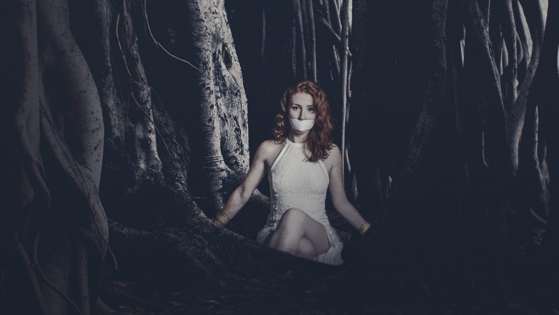 A woman with duct tape over her mouth sitting in a dark forest.