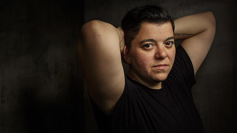 An image of Sinclair Sexsmith in a black t-shirt, looking at the camera with their arms cradled behind their head.
