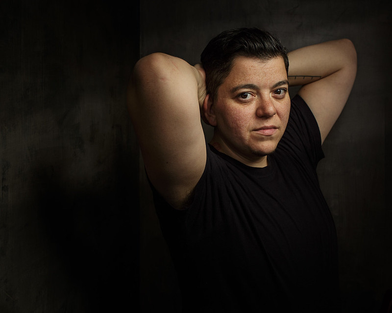 An image of Sinclair Sexsmith in a black t-shirt, looking at the camera with their arms cradled behind their head.